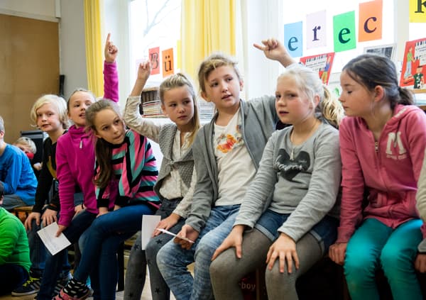 Viele Kinder sitzen nebeneinander auf einer Bank, ein paar melden sich und ein paar halten Zettel in der Hand.