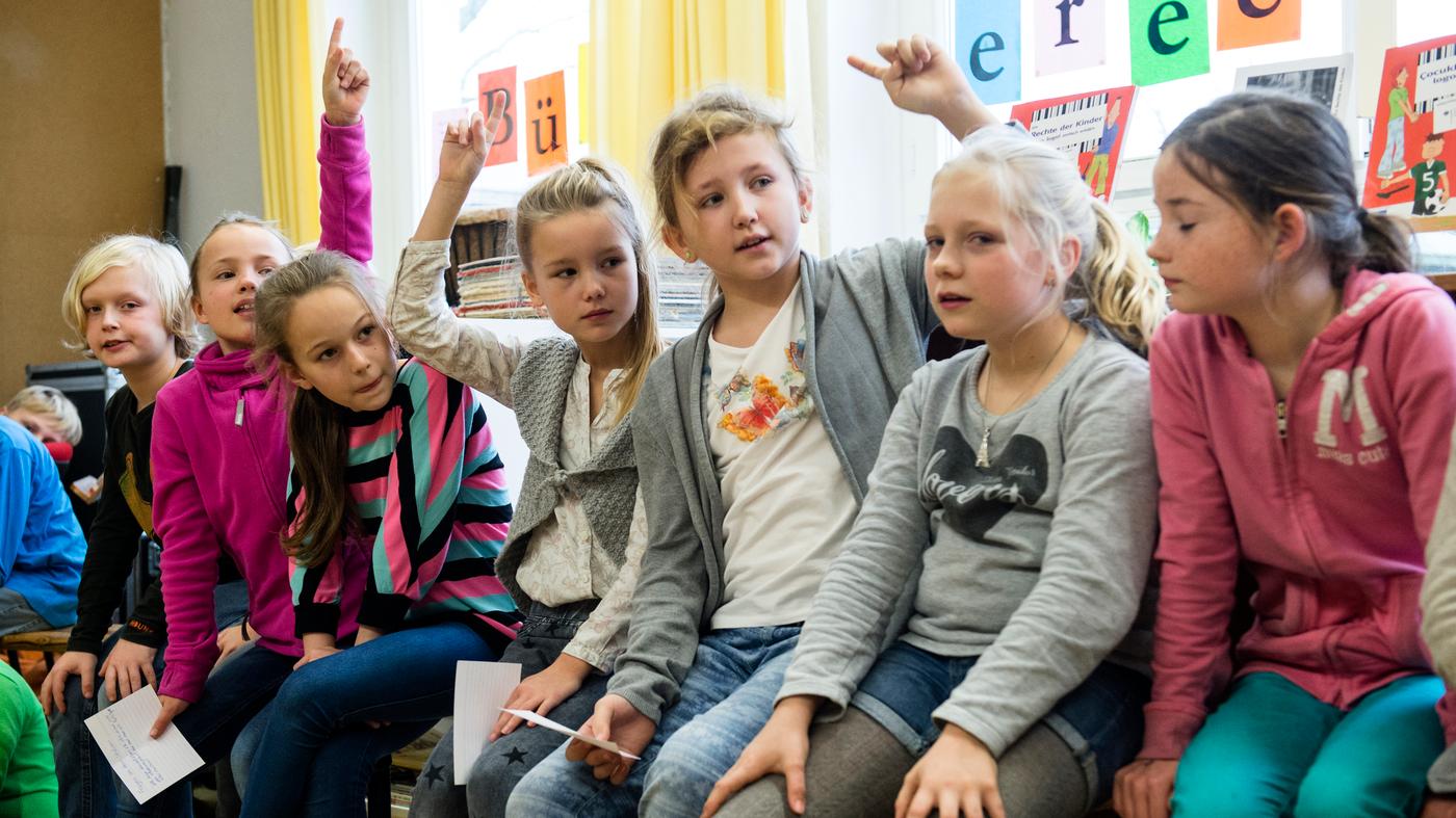 Viele Kinder sitzen nebeneinander auf einer Bank, ein paar melden sich und ein paar halten Zettel in der Hand.