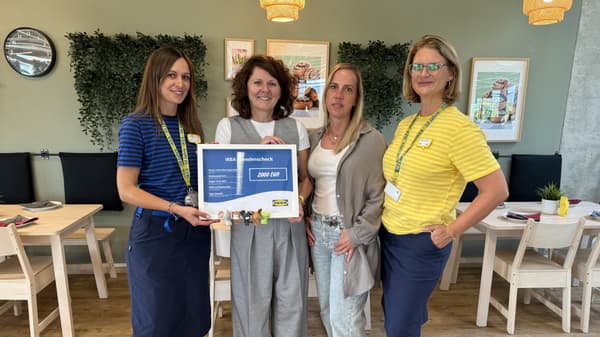 Vier Frauen stehen im IKEA Restaurant nebeneinander, im Hintergrund sind Holztische, Stühle, Pflanzen und Wanddeko zu sehen. Zwei der Frauen tragen IKEA-Mitarbeiterkleidung und überreichen einen Spendenscheck über 2.000 Euro an Vertreterinnen des Mutter-Vater-Kind-Hauses Aline, die Freizeitkleidung tragen. 