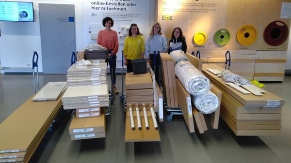 Vier Frauen stehen in einem IKEA-Markt hinter mehreren Transportwagen voller Möbelpakete, Teppiche und anderer Waren. Eine der Frauen trägt ein gelbes IKEA-Oberteil. Im Hintergrund befinden sich eine Recyclingstation und Infotafeln zum Online-Bestellen.  