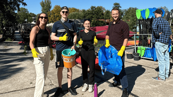 Vier junge Erwachsene mit gelben Handschuhen stehen bei einem Kajakverleih im Freien und halten Eimer, Müllzangen und eine blaue IKEA-Tasche. Sie lächeln und zeigen Daumen nach oben, während sie an einer Aufräumaktion teilnehmen.