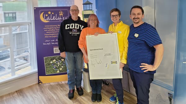 Vier lächelnde Personen stehen in einem Raum und präsentieren einen gerahmten Spendenscheck über 2500 € von IKEA. Im Hintergrund ist ein großer Banner des Vereins 