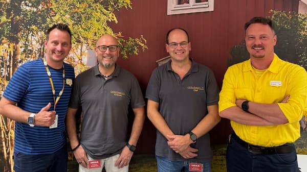 Vier Männer stehen vor einer Wand mit einem herbstlichen Naturmotiv. Zwei von ihnen tragen IKEA-Dienstkleidung, einer in Blau, der andere in Gelb. Die beiden anderen tragen graue Polohemden mit der Aufschrift „Lichtschmiede e.V.“. Alle lächeln und haben ihre Arme verschränkt oder zeigen eine positive Geste.  