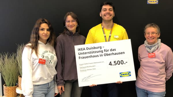 Vier Personen, darunter ein IKEA-Mitarbeiter in gelbem Shirt, halten einen Spendenscheck über 4'500 Euro für das Frauenhaus in Oberhausen. Sie stehen vor einer schwarzen Wand mit IKEA-Logo, links sind Dekorationspflanzen in Körben zu sehen.