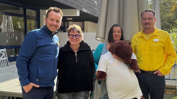 Vier Personen stehen im Aussenbereich eines Gebäudes zusammen. Zwei Männer tragen IKEA-Dienstkleidung, einer in Blau, der andere in Gelb. Eine Frau hält ein grosses Plüschtier in einem weissen T-Shirt. Alle blicken in die Kamera, einige lächeln. Im Hintergrund sind Tische, Stühle und eine grosse Sonnenschirmanlage zu sehen.  