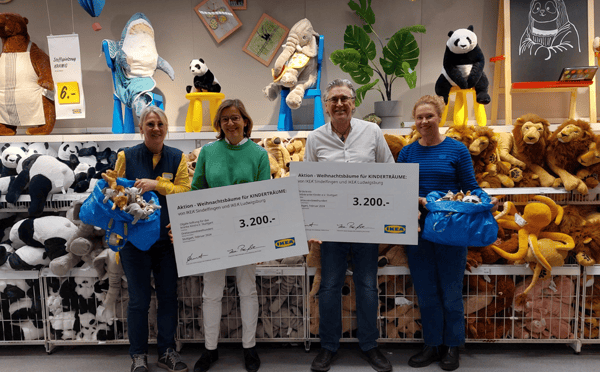 Vier Personen stehen in der Kuscheltier-Abteilung eines IKEA-Marktes vor Regalen voller Plüschtiere, darunter Löwen, Pandas und Oktopusse. Zwei von ihnen halten große Spendenschecks über jeweils 3.200 Euro mit der Aufschrift „Aktion – Weihnachtsbäume für KINDERTRÄUME“ von IKEA Sindelfingen und IKEA Ludwigsburg. Die beiden IKEA-Mitarbeitenden tragen blaue Westen mit gelben Shirts und halten blaue IKEA-Taschen voller Kuscheltiere.  