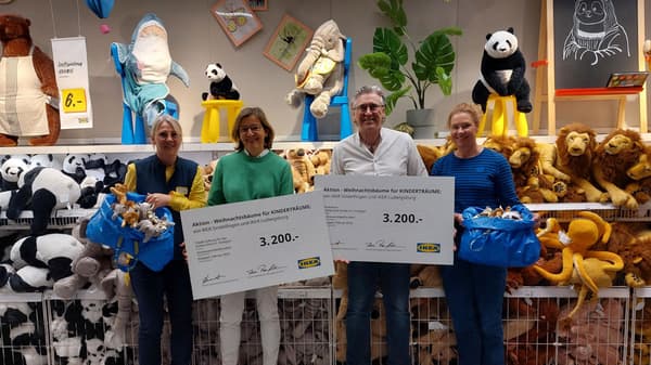Vier Personen stehen in der Kuscheltier-Abteilung eines IKEA-Marktes vor Regalen voller Plüschtiere, darunter Löwen, Pandas und Oktopusse. Zwei von ihnen halten große Spendenschecks über jeweils 3.200 Euro mit der Aufschrift „Aktion – Weihnachtsbäume für KINDERTRÄUME“ von IKEA Sindelfingen und IKEA Ludwigsburg. Die beiden IKEA-Mitarbeitenden tragen blaue Westen mit gelben Shirts und halten blaue IKEA-Taschen voller Kuscheltiere.  