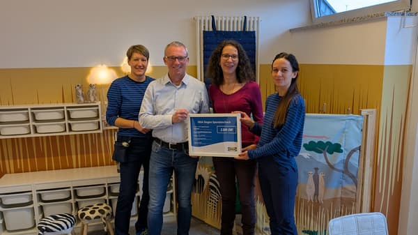 Vier Personen stehen in einem freundlich eingerichteten Kinderzimmer nebeneinander, im Hintergrund sind bunte Wanddekorationen, Spielzeugkisten und Kindermöbel zu sehen. Zwei Personen tragen IKEA-Mitarbeiterkleidung und überreichen einen Spendenscheck über 3.500 Euro an Vertreter:innen des Balthasar Kinder- und Jugendhospizes.