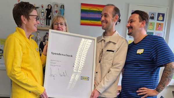 Vier Personen stehen in einem Innenraum und präsentieren gemeinsam einen gerahmten Spendenscheck von IKEA. Im Hintergrund sind Regale mit Puppen und eine Wanddekoration mit Regenbogenfarben zu erkennen.