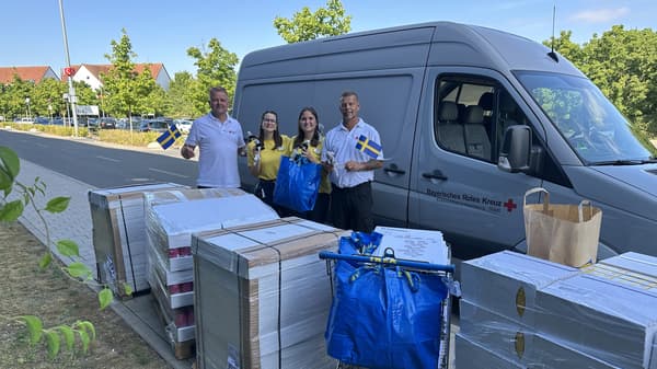 Vier Personen stehen neben einem grauen Transporter mit der Aufschrift: Bayerisches Rotes Kreuz. Auf dem Gehweg daneben befinden sich mehrere große, in Folie verpackte Paletten und blaue IKEA-Taschen mit Gegenständen. Im Hintergrund sind Bäume und Gebäude zu sehen.
