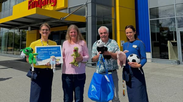 Vier Personen stehen vor dem Eingang eines IKEA-Marktes in Siegen und halten einen Spendenscheck über 2.000 Euro sowie mehrere Kuscheltiere. Zwei der Personen tragen IKEA-Mitarbeiterkleidung, während die anderen beiden in Freizeitkleidung gekleidet sind.  