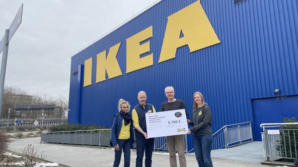 Vier Personen stehen vor der blauen Fassade eines IKEA-Gebäudes mit großem gelben Logo und halten gemeinsam einen Spendenscheck über 5.750 Euro. Zwei der Personen tragen IKEA-Mitarbeiterkleidung mit gelben und blauen Elementen, während die anderen beiden dunklere Kleidung mit einem grünen Logo tragen. Der Scheck trägt den Schriftzug „Dein IKEA Weihnachtsbaum erfüllt einen Kindertraum“.  