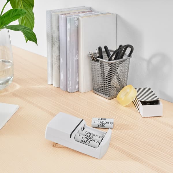 Vier weiße IKEA LADDA Batterien liegen auf einem Schreibtisch in hellem Holz, zwei davon stecken in einem Aufladegerät und zwei daneben.