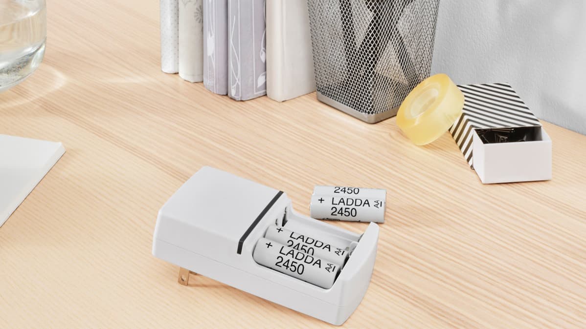 Vier weiße IKEA LADDA Batterien liegen auf einem Schreibtisch in hellem Holz, zwei davon stecken in einem Aufladegerät und zwei daneben.