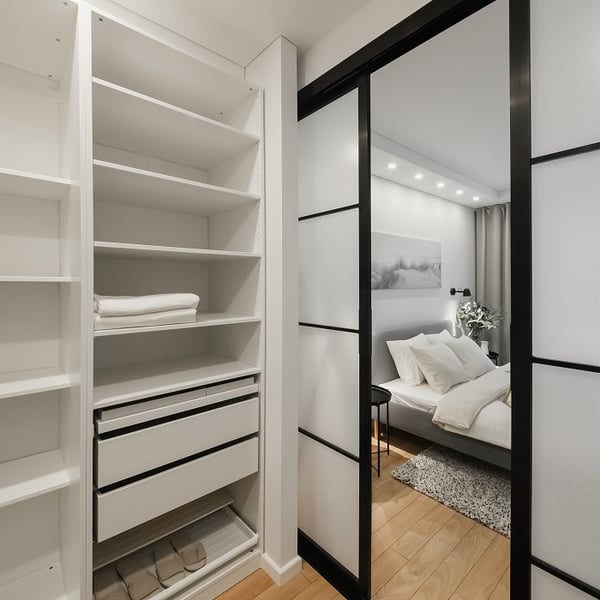 View from a walk-in closet into a cozy, modern bedroom through black-framed sliding glass doors. The closet features white open shelving with folded linens, organized drawers, and storage boxes on the bottom shelf. The bedroom beyond has a neatly made bed with soft pillows, ambient ceiling lighting, and minimalist decor.