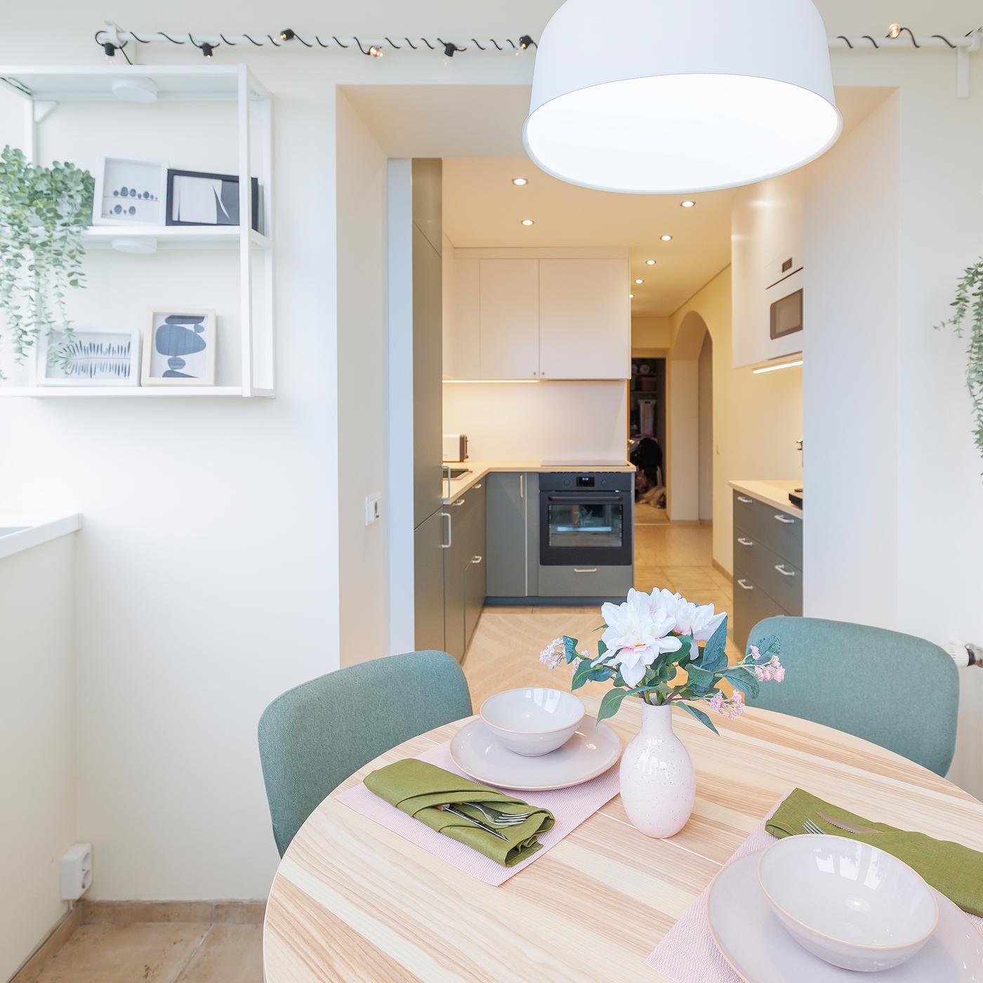 View from the dining area into the kitchen, showing a set table with soft pastel tableware, green napkins and a pendant lamp overhead.