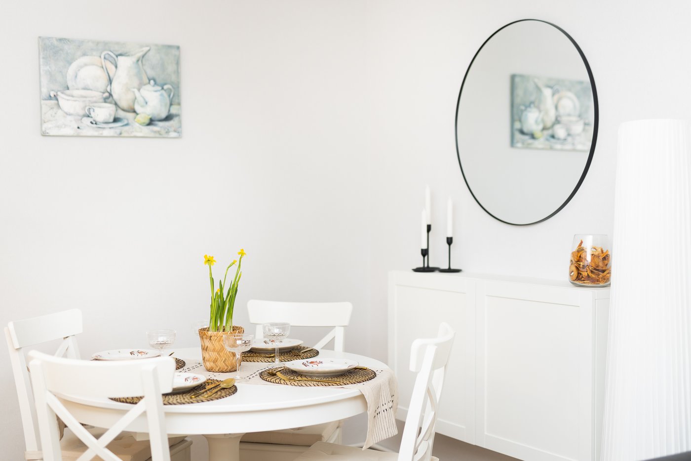 View of a dining area with a set table, a white sideboard, and a round wall mirror. The space appears bright, tidy, and cosy.