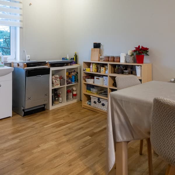 View of the dining area with a table and chairs, and a children’s toy shelf in the background; a bright room with a window and wooden flooring.