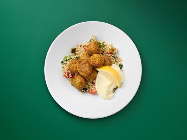 Vihreää taustaa vasten falafelannos, jossa falafel pyöryköitä, couscousia, kasviksia ja aiolia sitruunaviipaleen kera.