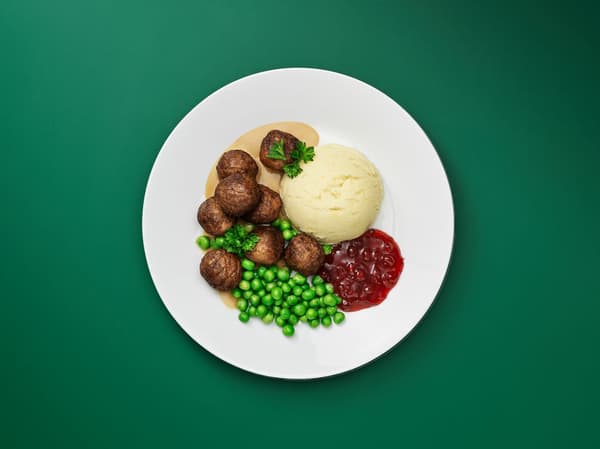 Vihreää taustaa vasten pyörykkäannos, jossa lihapyöryköitä, perunamuusia, herneitä ja puolukkahilloa.