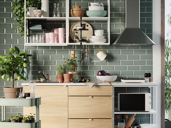 Vihreäksi laatoitettu keittiö, avohyllyillä asitoita, liesituuletin, yrttejä pöydällä ja pieni sitruunapuu.