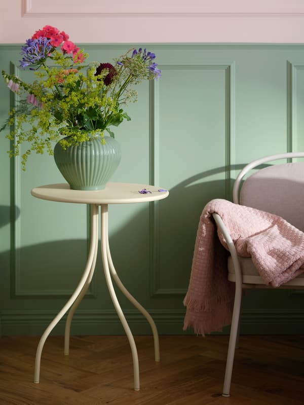 Világossárga TÅNEBRO dohányzóasztal áll egy világosszürke-bézs ISMANTORP fotel mellett, a fotel karfáján pedig egy világosrózsaszín HORNMAL takaró látható.