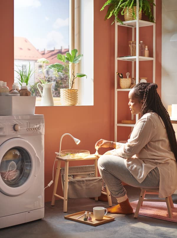 ผู้หญิงนั่งอยู่ข้างหน้าต่างที่มีแสงแดดส่องบนเก้าอี้เตี้ย VILTO/วิลโต ที่มีชุดดูแลเล็บวางอยู่บนสตูลเก็บของ VILTO/วิลโต ข้างเครื่องซักผ้า