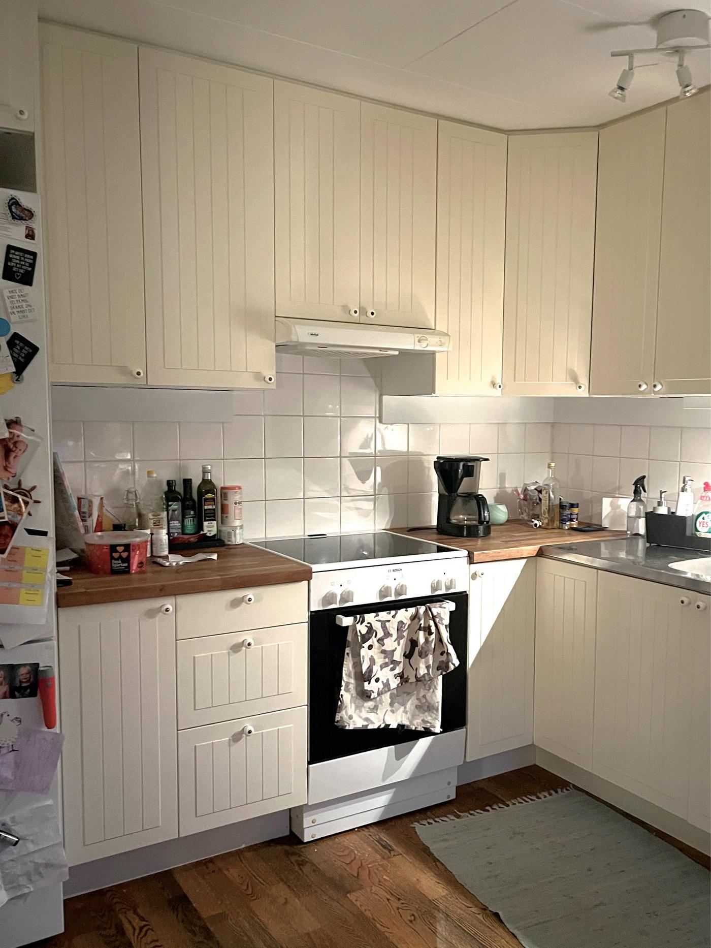 Vinkelkök med ljusa luckor, vit spis och vitt kakel. Solljus faller in över bänken där hushållsprodukter och en kaffemaskin står framme