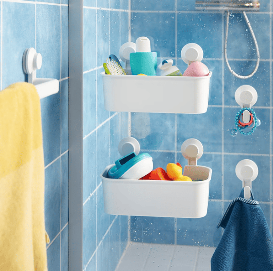 Vista de cerca de canastos plásticos TISKEN afirmados con ventosas a la mampara de baño. Se ven también perchas y un toallero TISKEN con una toalla amarilla colgada. Los muros del baño están revestidos en cerámicas celestes cuadradas con fragüe blanco. 