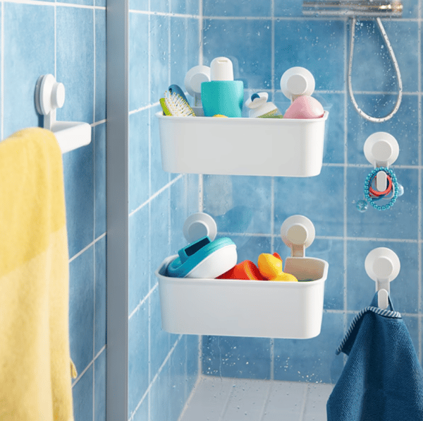 Vista de cerca de canastos plásticos TISKEN afirmados con ventosas a la mampara de baño. Se ven también perchas y un toallero TISKEN con una toalla amarilla colgada. Los muros del baño están revestidos en cerámicas celestes cuadradas con fragüe blanco. 