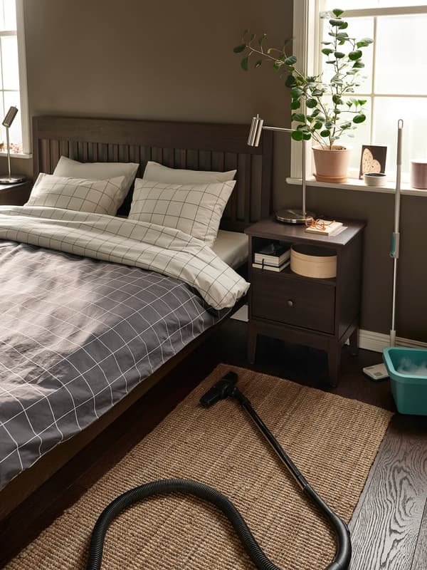 Vista de una habitación con una cama y un velador. Al costado de la cama se ve una aspiradora sobre una alfombra.