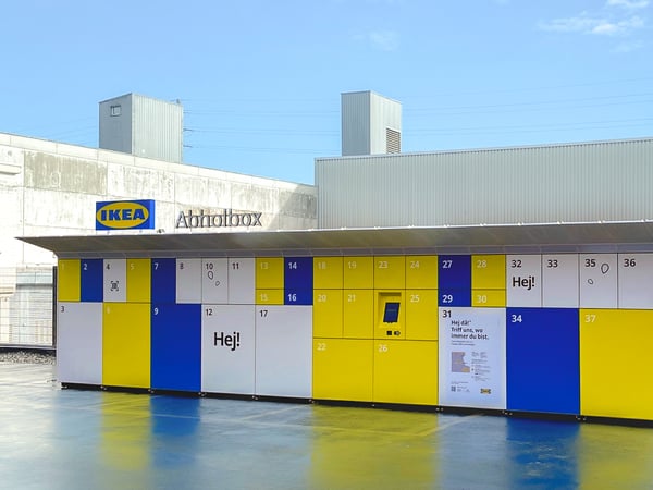 Vista di un locker IKEA esterno: un grande container con diversi scomparti numerati e di varie dimensioni nei colori bianco, giallo e blu e con il logo IKEA sul tetto, accanto al quale è apposta la scritta «IKEA locker»