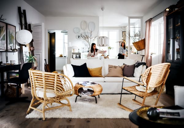 Vista frontal de un living con un sofá blanco BACKSÄLEN de tres cuerpos con cojines sobre el. En frente hay una mesa de centro con espacio para almacenamiento en su interior y dos sillas de madera.