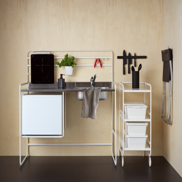Vista frontal de una mini cocina SUNNERSTA con lavaplatos en acero inoxidable incorporado, encimera metálica y estructura blanca. La mini cocina está situada frente a un muro color madera y sobre un piso de color negro. 