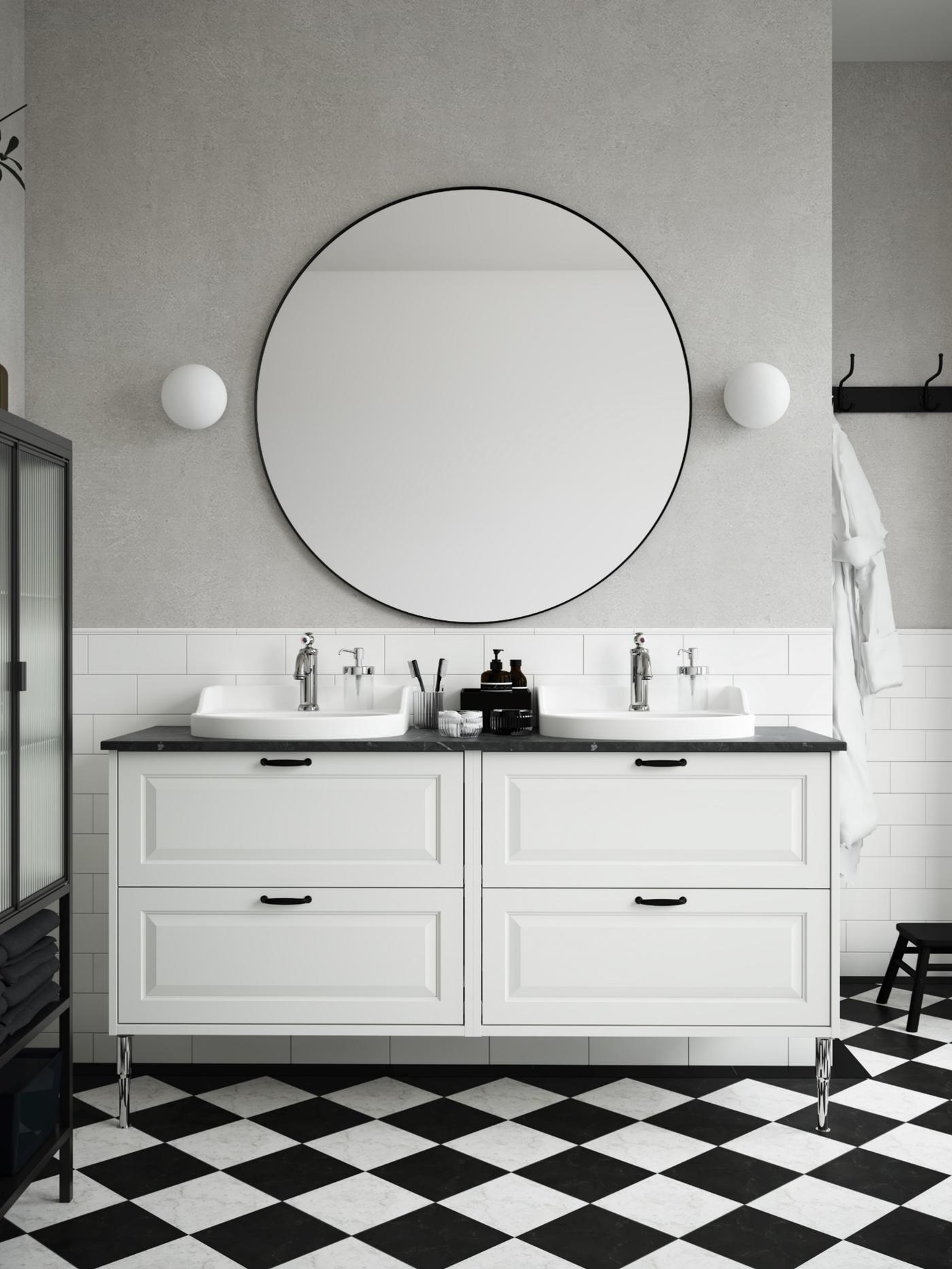Vista frontale di un bagno TÄNNFORSEN bianco composto da un mobile per lavabo a quattro cassetti bianco, uno specchio grande rotondo e due lampade da parete a sfera bianche. Il pavimento è a scacchi bianco e nero