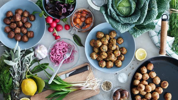 Vista superior de una mesa con platos celestes y negros que contienen albóndigas IKEA, un plato con cebolla morada, una tabla para picar de madera con un cebollín y otras verduras listas para prepararse. 
