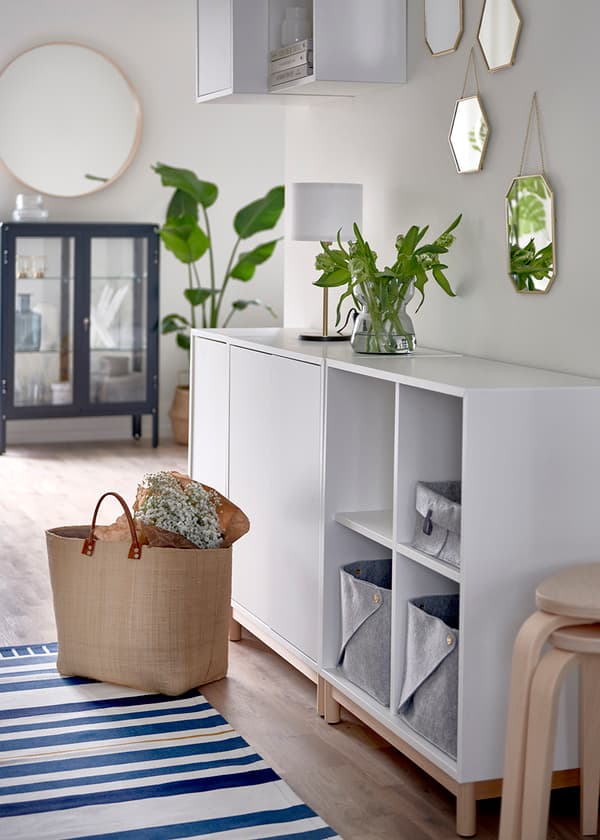 Vit EKET förvaringsmöblel med fyra öppna fack och tre skåp mot en ljus vägg. Framför, på en blå- och vitrandig matta, står en beige väska med en bukett med brudslöja inlaget i brunt papper. 