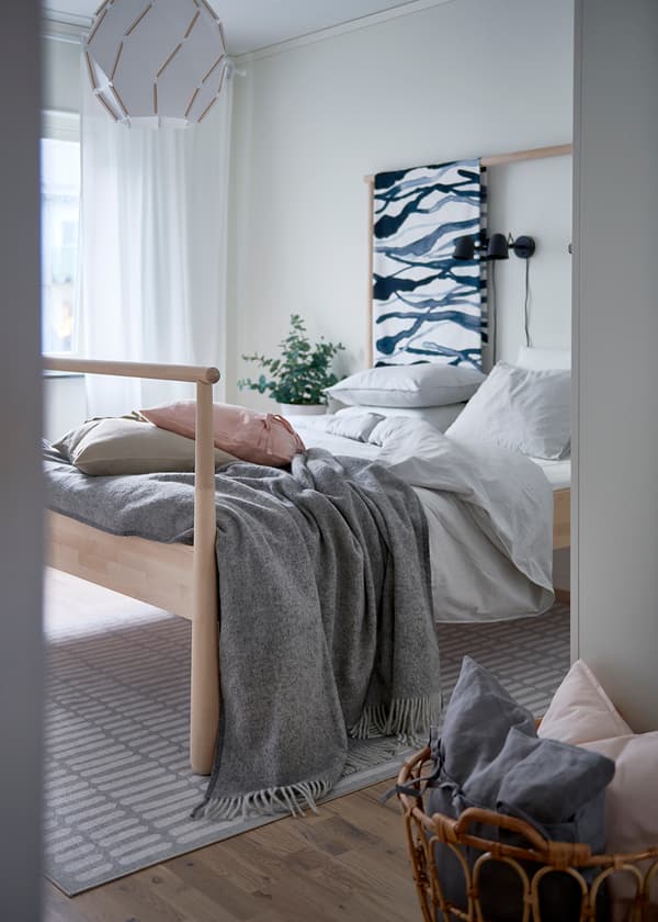 Vitt sovrum med en BJÖRKSTA säng bäddad med ljusgrå påslakan och en ljusgrå filt. I förgrunden skymtas en SNIDAD korg innehållandes två kuddar. 