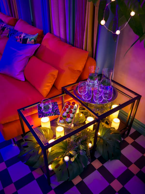 VITTSJÖ egymásba rakható asztalok. Nassolnivalókkal, FEJKA szőnyeggel és SMYCKA műnövényekkel, illetve GODAFTON LED tömbgyertyákkal.