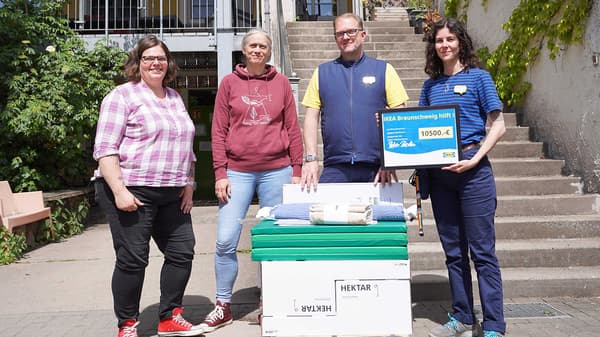 Vor der Treppe zur Wohngruppe Ohe stehen 4 Personen. 2 Mitarbeitende der ESN und 2 IKEA Mitarbeitende. Eine Person zeigt das Schild mit der Spendensumme. Vor der Gruppe steht ein Stapel mit Wohnaccessoires.