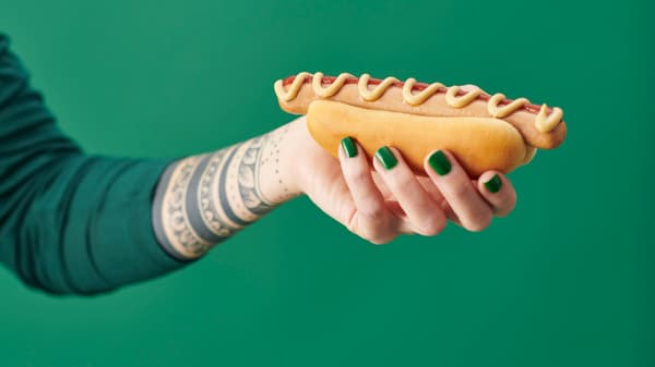 Vor einem grünen Hintergrund hält eine Frau den neuen PLANT-HOTDOG von IKEA in der Hand.