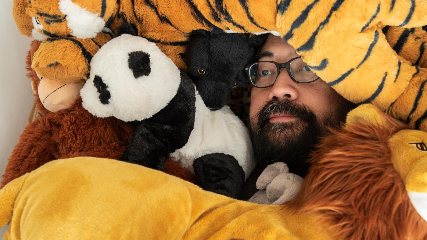 Vousatý muž v brýlích leží uprostřed hromady plyšových hraček DJUNGELSKOG. Vidět je mezi nimi jen jeho tvář.