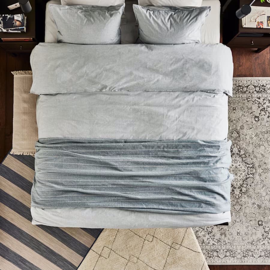 Vrchní pohled na postel, na které je ustlaná šedá přikrývka, přehoz a polštáře. Pod postelí jsou 4 různé koberce béžových barev a různých vzorů.