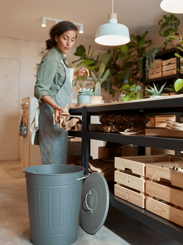 Vrouw staat in een kamer vol planten en gooit afval in een vuilnisbak.