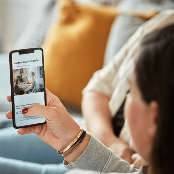 Vrouw zit op een bank en heeft een telefoon in haar handen met de IKEA app geopend. Op het scherm is ze een IKEA Family profiel aan het aanmaken.