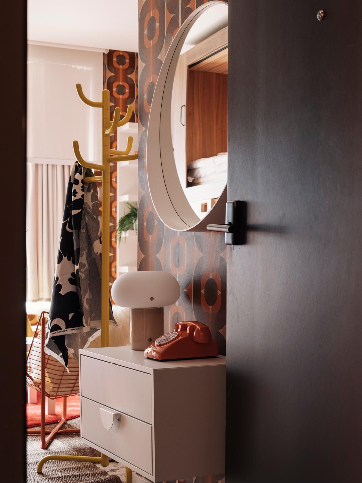 Vue à travers une porte ouverte sur une pièce rétro avec du papier peint géométrique, une commode blanche avec un téléphone vintage orange et une lampe blanche, un porte-manteau jaune avec des manteaux, des rideaux, des plantes et une chaise en arrière-plan.