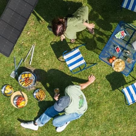 Vue aérienne de deux personnes en train de pique-niquer dans un pré. Autour d'elles se trouvent des chaises, une table, un parasol et des panneaux solaires portables.