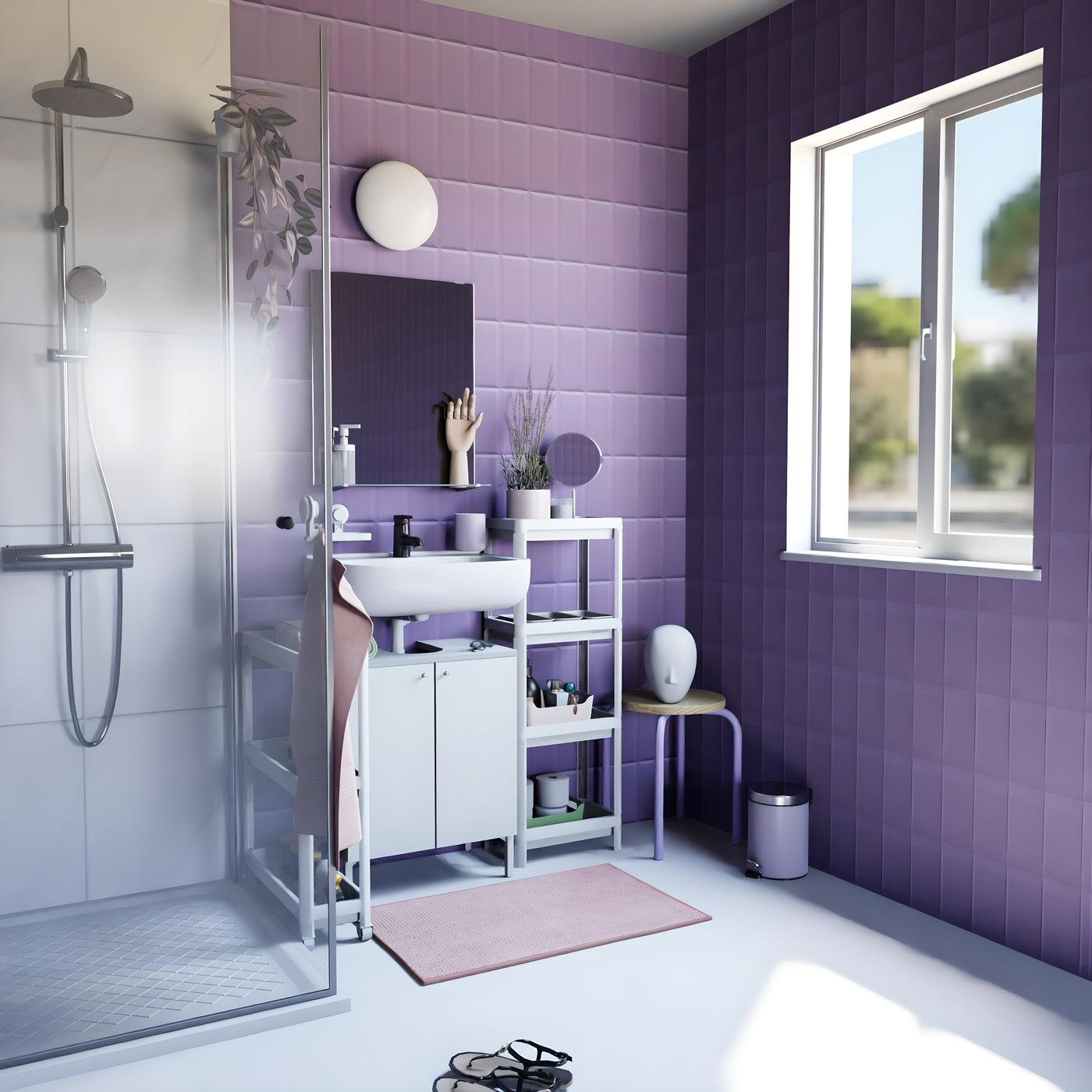 Vue de biais d'une salle de bain aux carrelages mauves, avec une douche et du mobilier blanc et mauve. Accessoires mauves et linges de bain roses