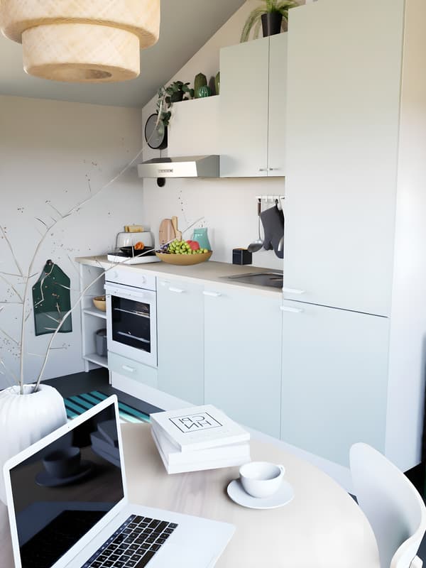 Vue de côté d'une cuisine verte claire sous pente, avec plan de travail en bois, sol noir et linge de cuisine vert. Abat-jour en osier, et table à manger ronde au premier plan, avec ordinateur et décorations.