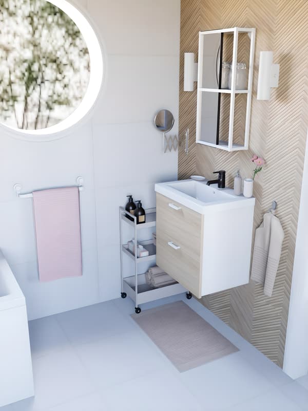 vue de côté d'une salle de bain blanche et bois effet chene, dans une suite parentale, avec du linge de bain rose et beige, un mur de carrelage imitation chevron bois clair, et une baignoire blanche. Fenêtre ronde montrant un extérieur 